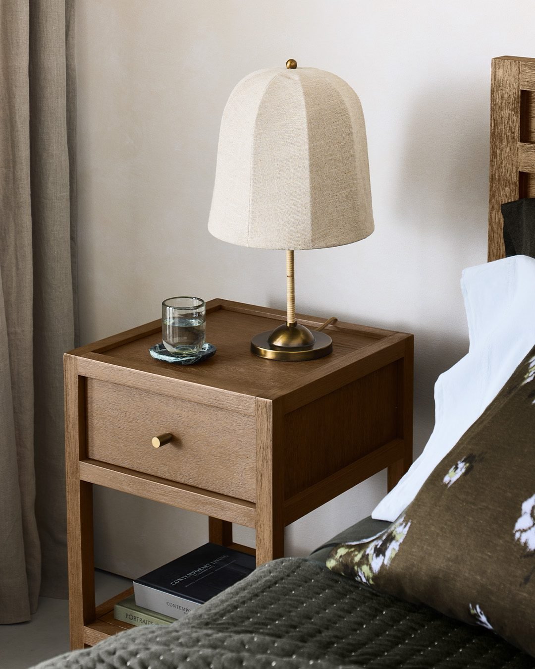 A simple, light oak nightstand with one drawer and an open lower shelf. On the top sits a water glass on a blue stone coaster and a unique lamp with a brass base and a textured fabric shade shaped like a bell.