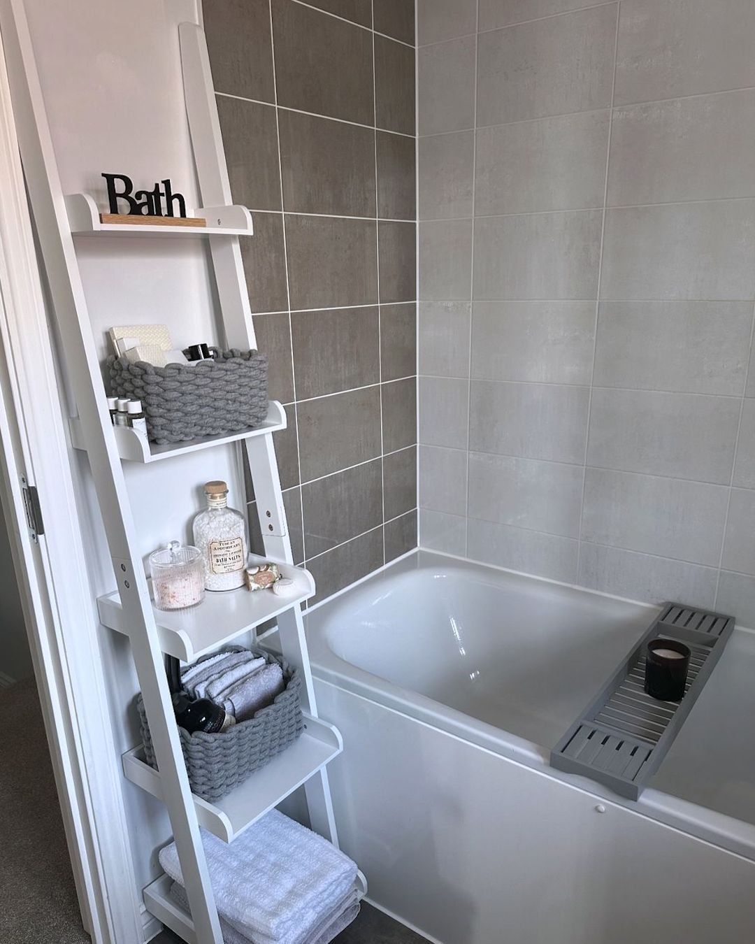 Tall white leaning ladder shelving unit next to a bathtub, storing rolled white towels, gray woven baskets, bath salts, and decorative accessories.