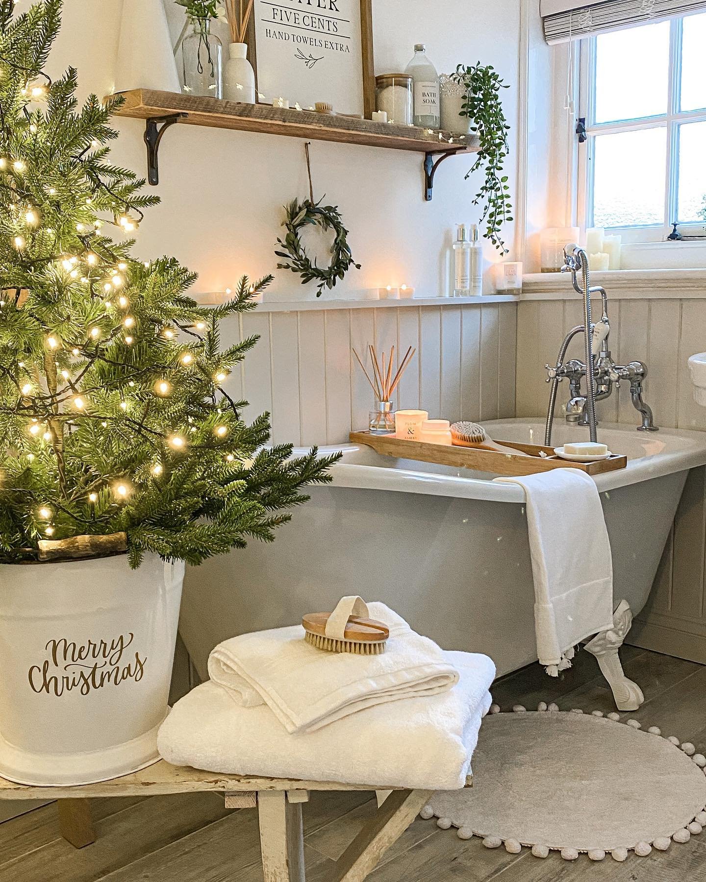 A cozy bathroom with a clawfoot tub, featuring a large, brightly lit Christmas tree in a "Merry Christmas" bucket and candles creating a soft glow.