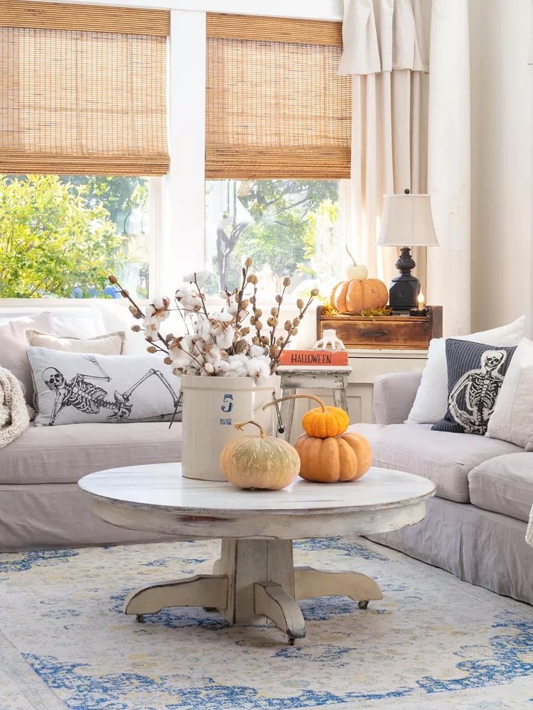 Farmhouse living room with a distressed white coffee table, skeleton pillows on a light gray sofa, and pumpkins and cotton stems as decor.