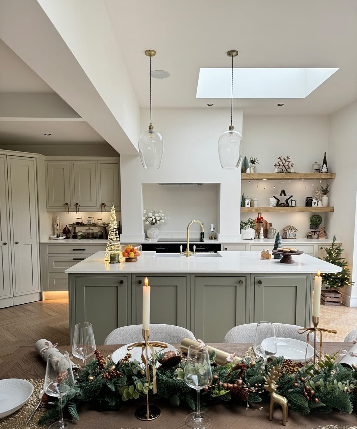 A modern, light-filled kitchen featuring a large island with a soft sage green base and white counter. The adjacent dining table is set with a beautiful holiday tablescape using a thick pine garland, gold candlesticks, and reindeer figures.