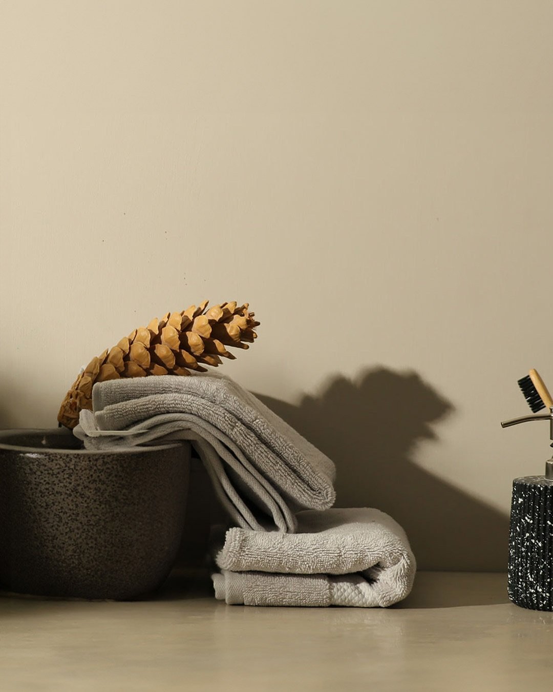 Small stack of light grey hand towels next to a dark grey textured bowl and a large decorative pine cone.