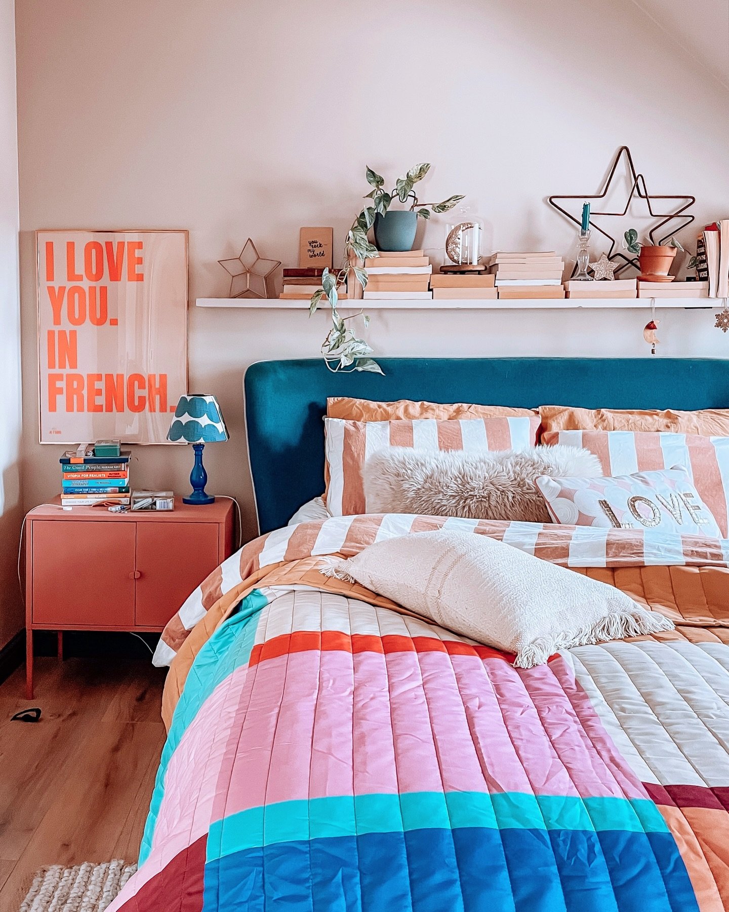 A colorful and eclectic bedroom with a bed featuring a multi-colored patchwork quilt, a dark teal headboard, a coral-pink nightstand, and a gallery shelf above the bed.