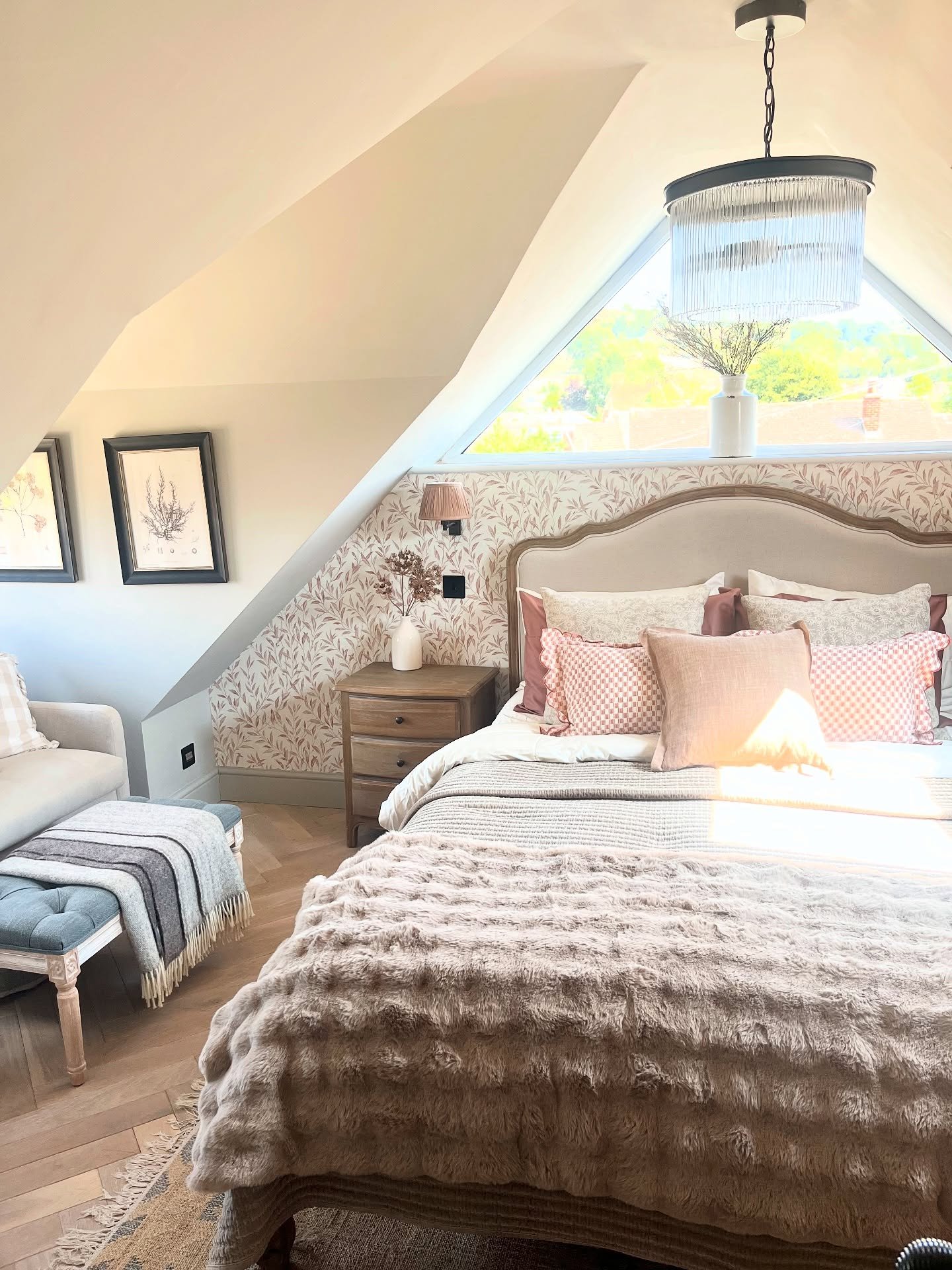 A cozy loft bedroom with a large triangular window, a bed with an arched headboard, and a soft pink leaf-patterned wallpaper accent wall.