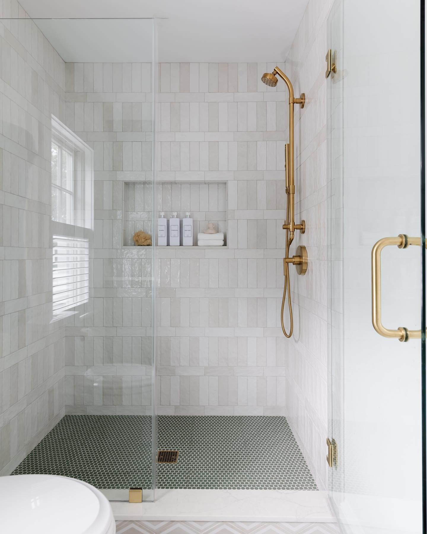 A bright walk-in shower with vertical, slightly varied white subway tiles, a floor of dark green penny tile, and polished brass fixtures.
