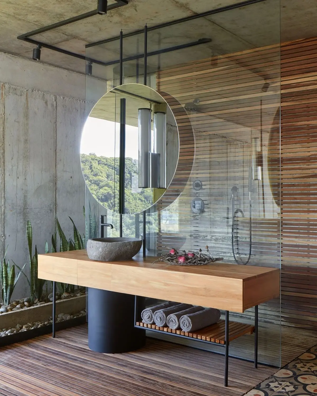 A bathroom vanity with a long, light wood countertop resting on a black metal frame and a cylindrical black pedestal base. A rough-hewn, gray stone vessel sink sits on the countertop, paired with a black faucet.