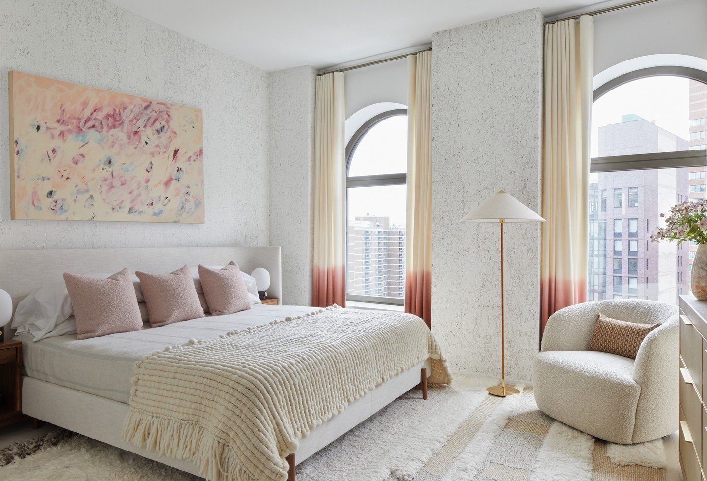 A cozy bedroom with textured white walls, a plush bed with soft pink pillows, and tall arched windows with dip-dyed curtains.