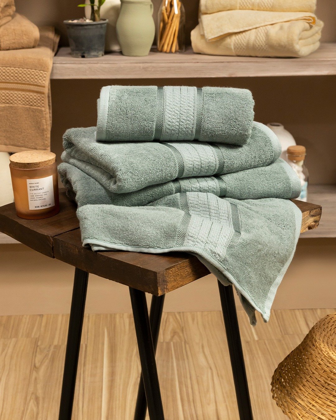Stack of plush sage green bath towels with patterned borders, displayed on a dark wood and metal stool.