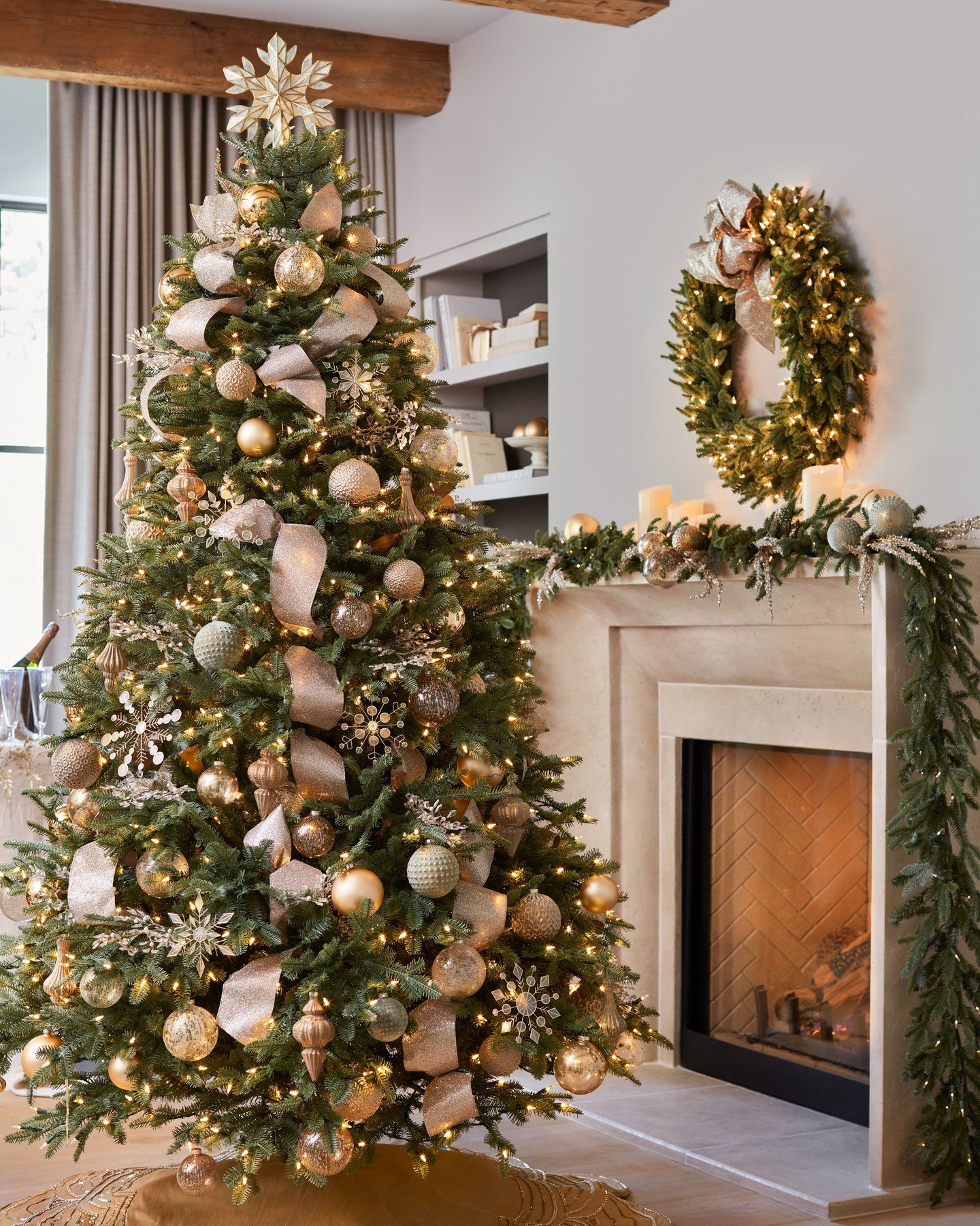 A Christmas tree decorated with a wide, metallic champagne ribbon, gold and pale green ornaments, and sparkling snowflake picks next to a fireplace.