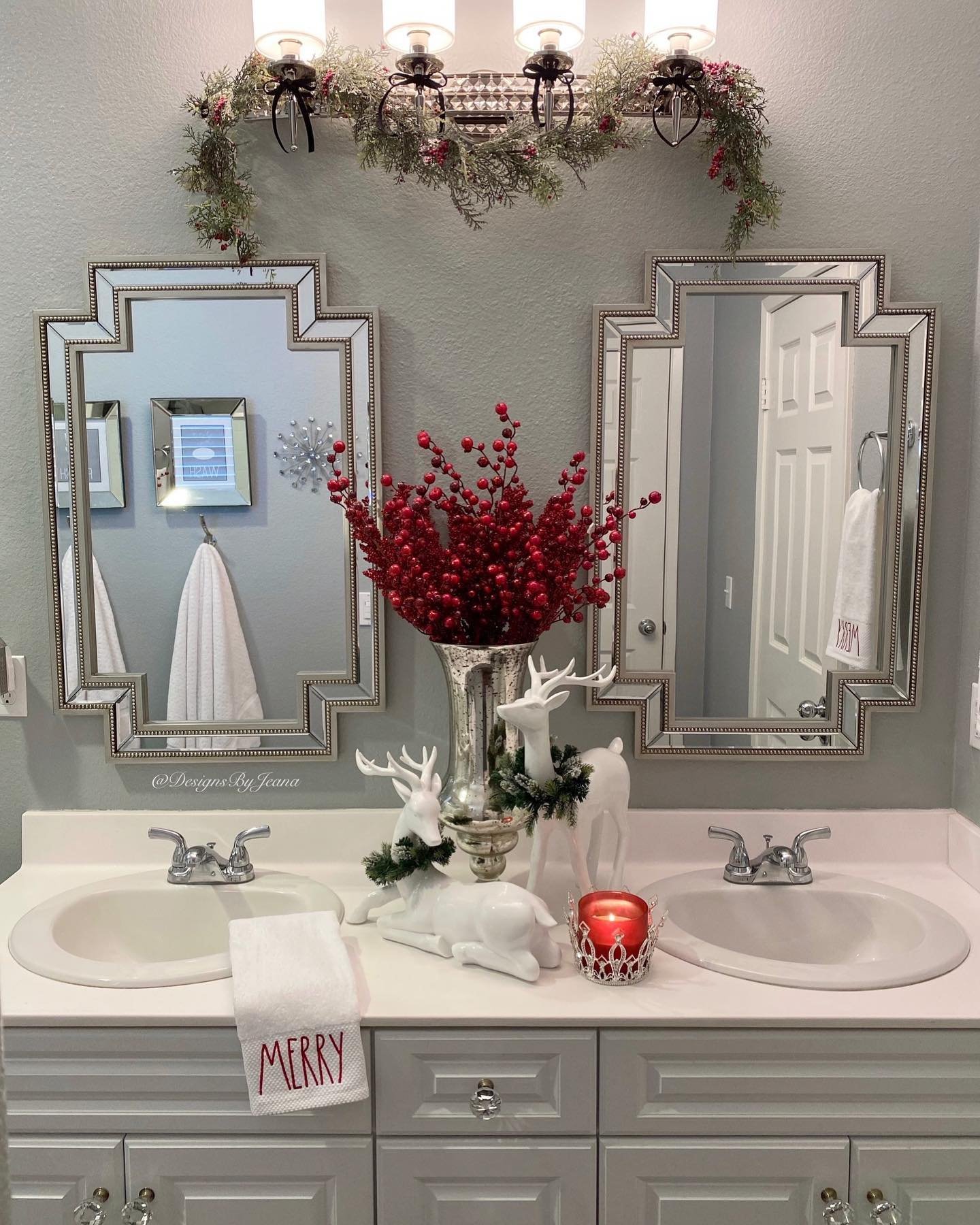 A double vanity decorated for Christmas with a large vase of red berries, white ceramic deer figures, and a garland draped over the light fixture.