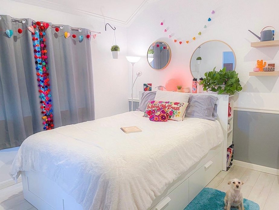 A playful bedroom with a white bed, colorful pom-pom and heart garlands, two round mirrors above the bed, and a small dog sitting on a blue rug.