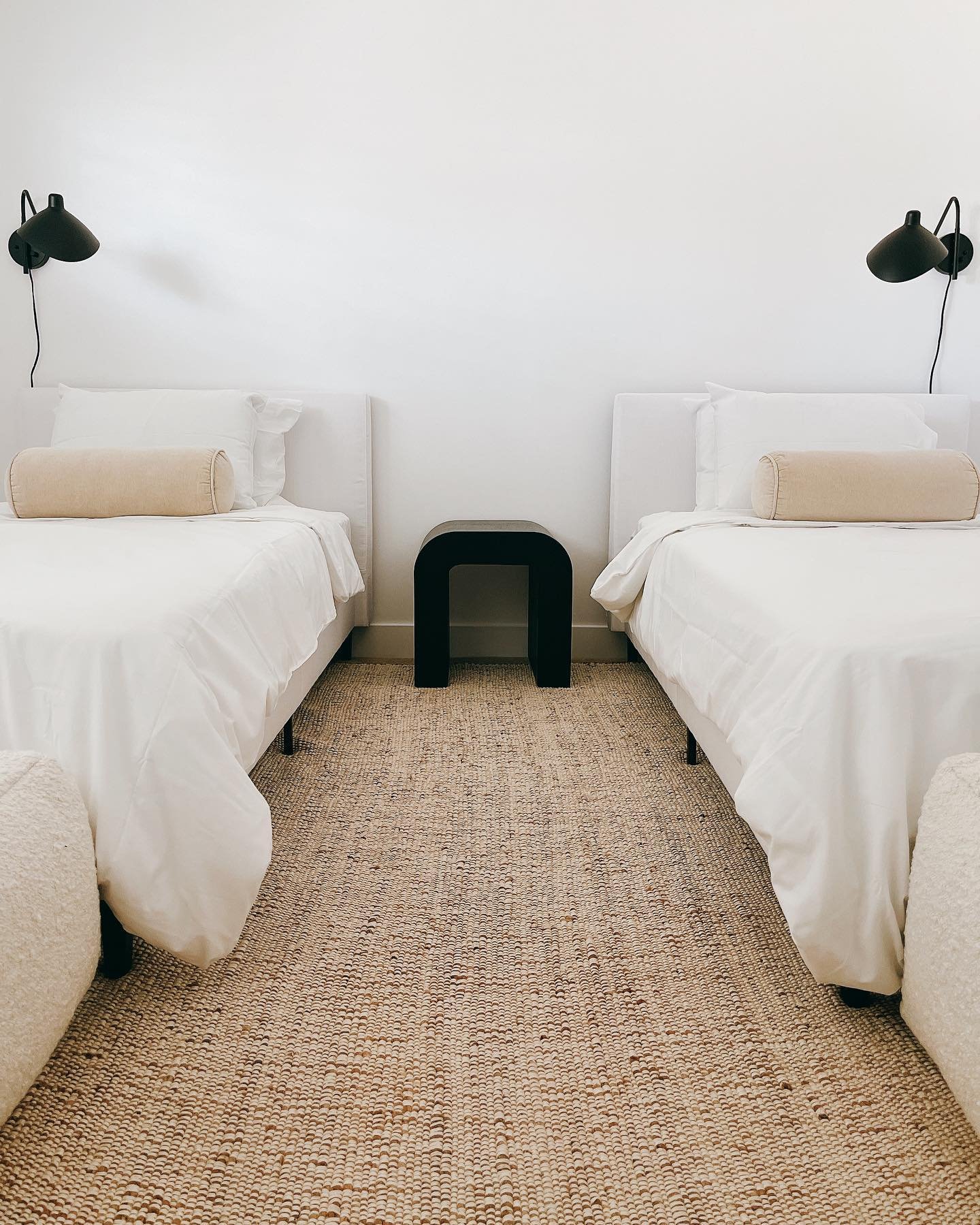 A minimalist twin bedroom with two white beds, a black U-shaped nightstand, and black wall sconces.