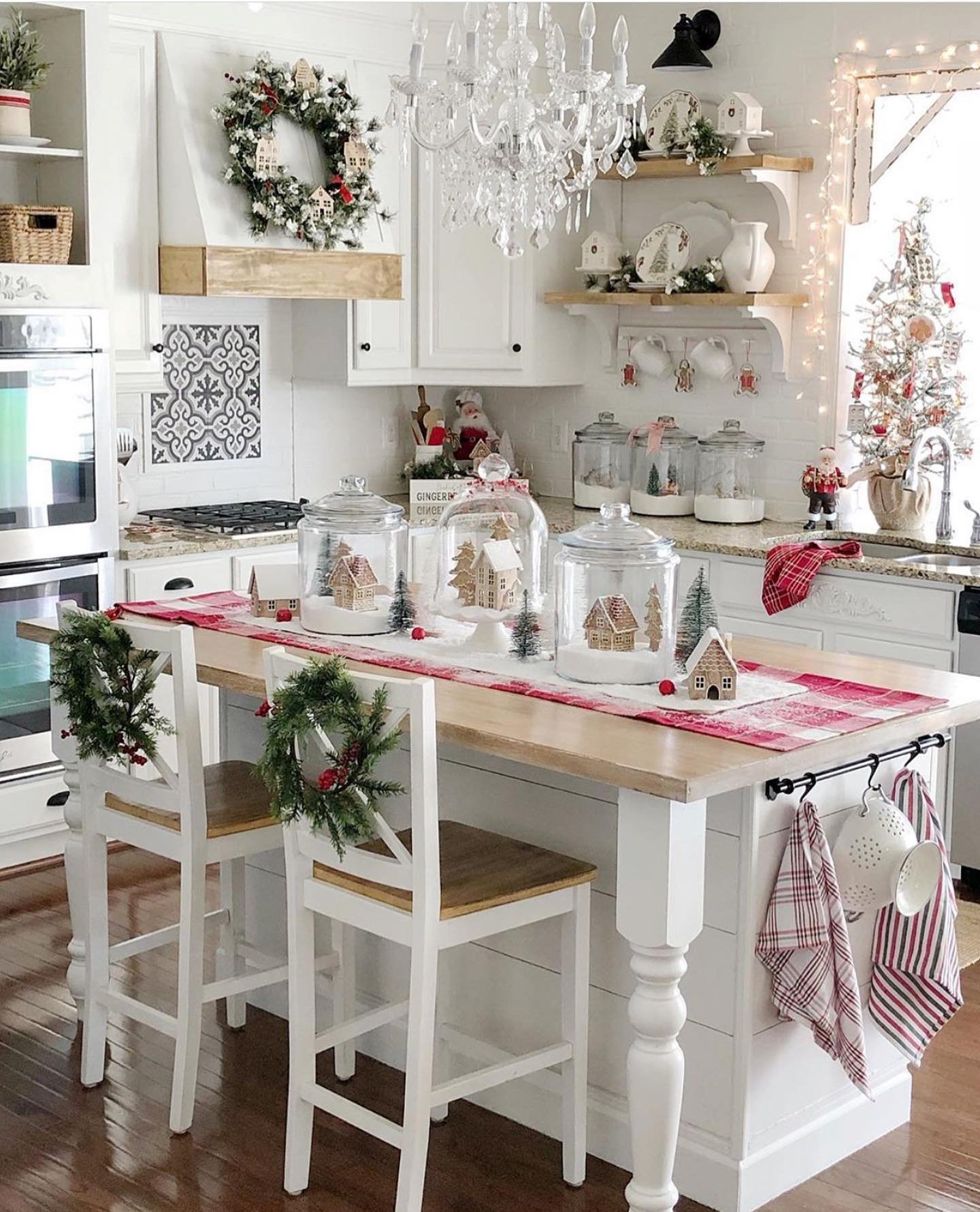 A cozy white farmhouse kitchen decorated for Christmas, featuring a large island topped with a red and white plaid runner and three glass jars containing miniature gingerbread house scenes. A crystal chandelier hangs above.