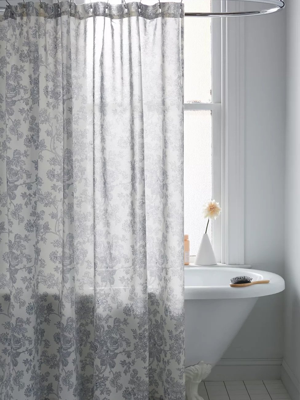 A semi-sheer white shower curtain with a dense, repeating floral and foliage pattern in a muted light gray or silver tone.