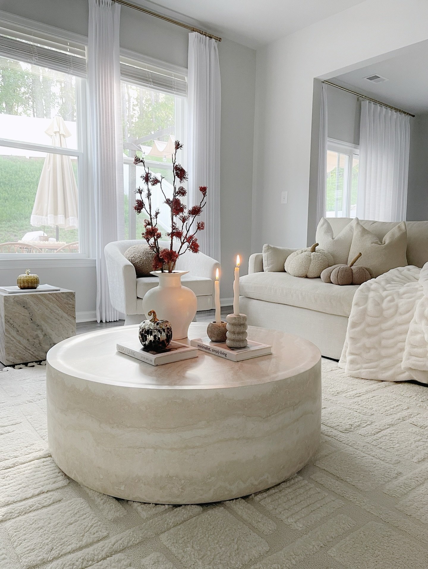 Modern living room with a large, round travertine coffee table, a cream-colored sofa, and minimalist fall decor.