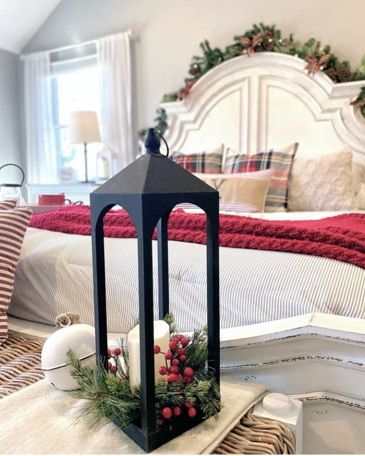 A close-up view of a black metal lantern holding a candle, pine sprigs, and red berries, with a bed in the background dressed in red knit and striped bedding, and a distressed white headboard decorated with garland.