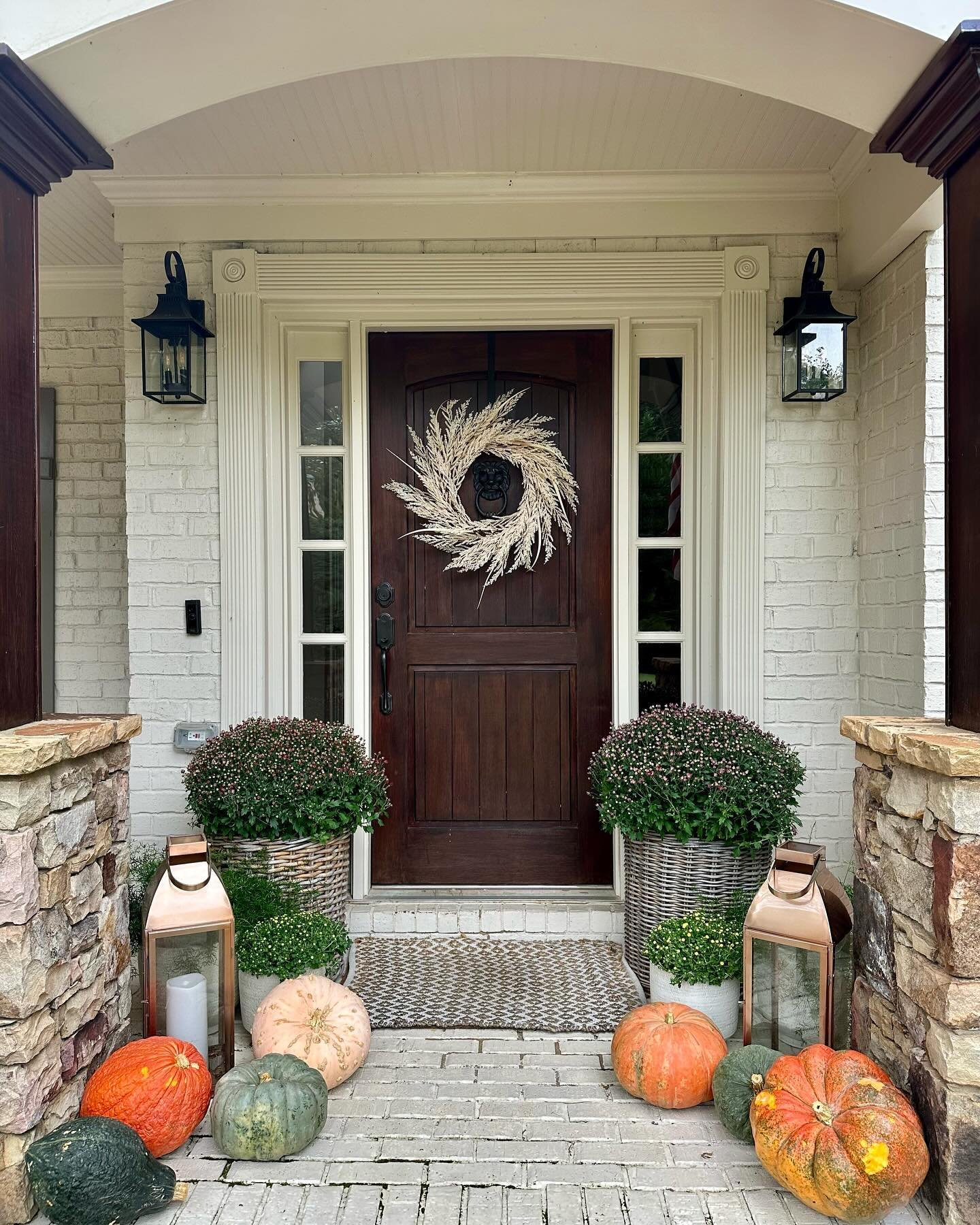A beautiful front porch with a dark wood door, decorated with pumpkins, large planters of mums in woven baskets, and rustic lanterns.