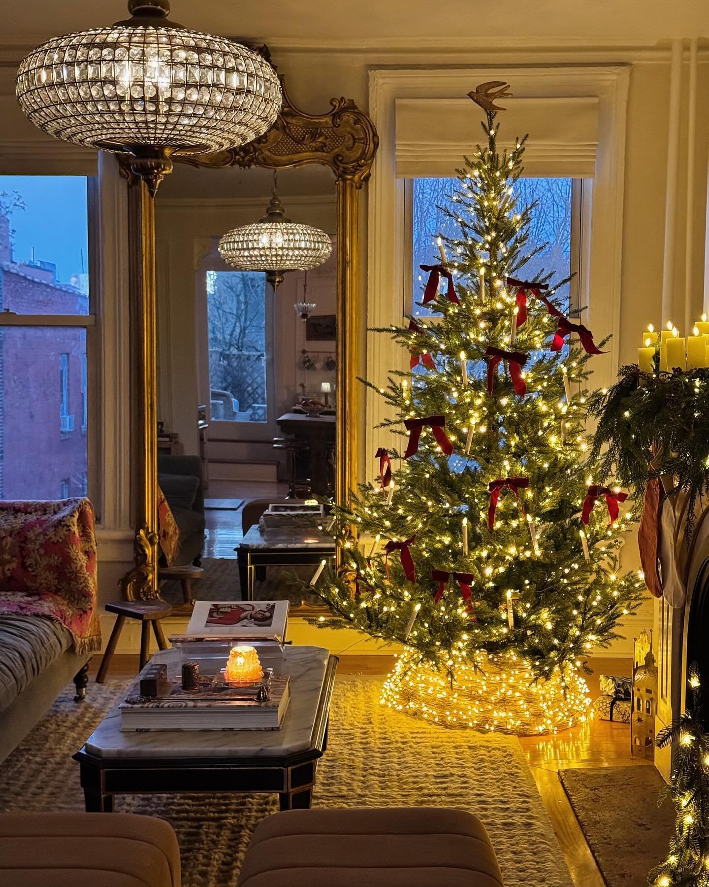 Dark and elegant living room featuring a brightly lit Christmas tree with red bows, and a large gold ornate mirror.