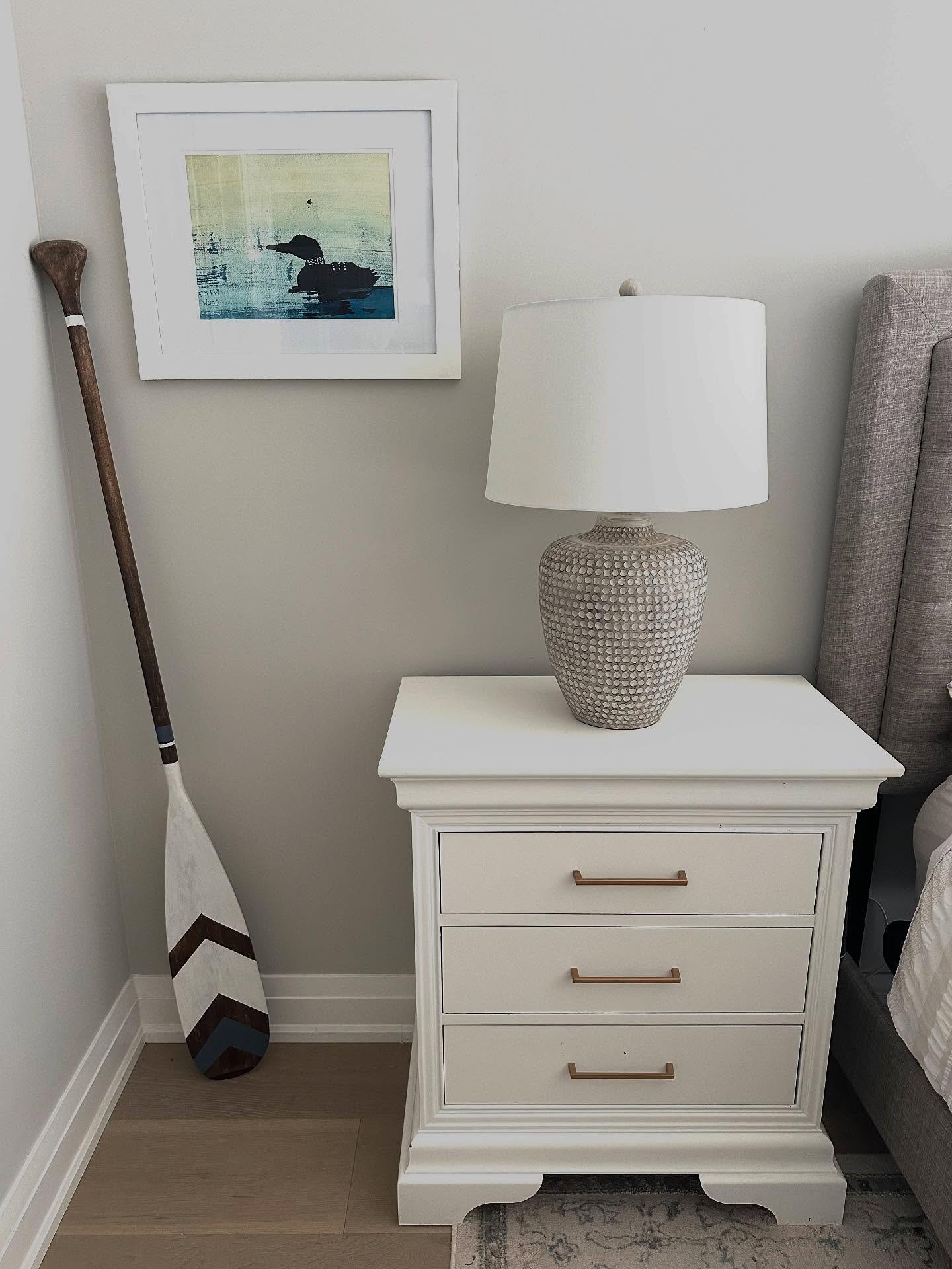 A white, three-drawer nightstand with bronze bar pulls and a decorative base. A large, textured ceramic lamp with a white shade sits on top. A framed picture of a loon is on the wall, and a wooden paddle leans in the corner.