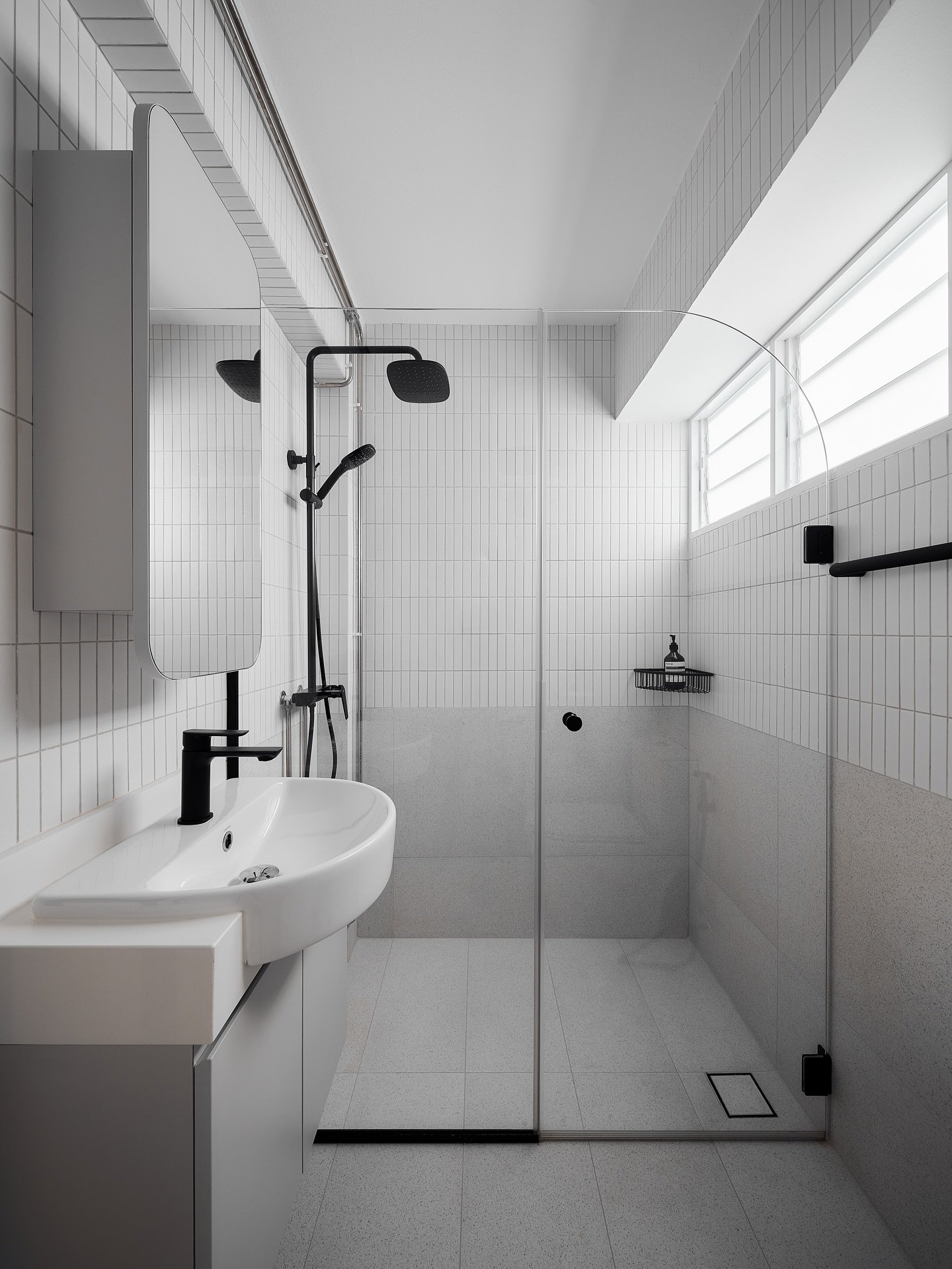 A minimalist black and white bathroom with a two-tone tiled wall. The upper half features white vertical rectangular tiles and the lower half has speckled gray-beige square tiles.