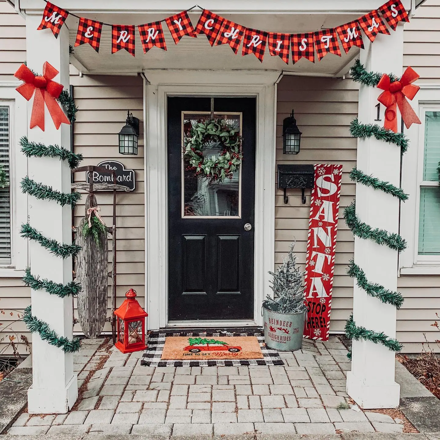 A cozy Christmas porch with white columns wrapped in thin green garland, a "MERRY CHRISTMAS" banner in red and black buffalo plaid, and a vertical "SANTA STOP HERE" sign.
