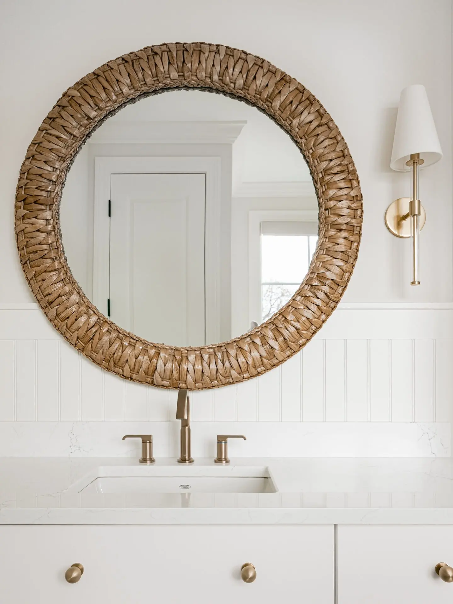 A bathroom with a large, round mirror with a thick, woven rattan frame over a white vanity.