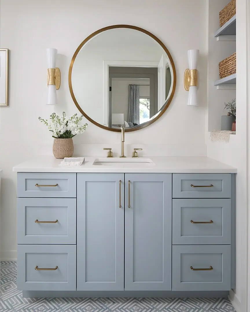 A light blue bathroom vanity with multiple drawers and a central cabinet, topped with a white countertop and an undermount sink. It's paired with a large round mirror with a gold frame and two elegant gold-and-white sconces, against a white wall.