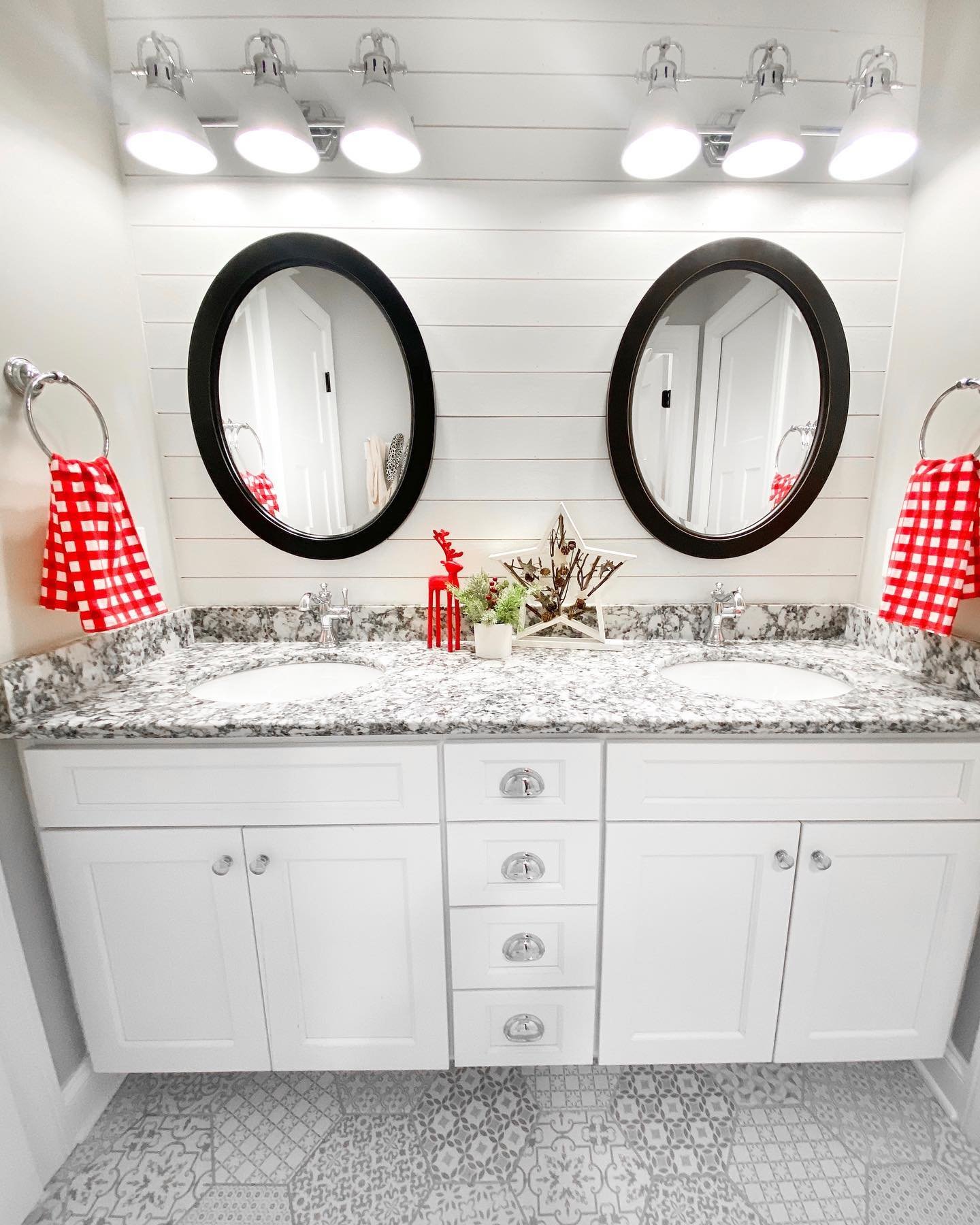  A white double vanity with shiplap walls, black oval mirrors, and red and white gingham towels on either side.