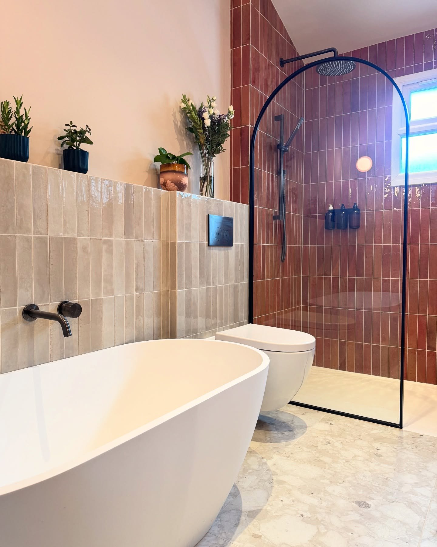 A colorful modern bathroom featuring a walk-in shower with deep terracotta/red vertical subway tiles and a black-framed arched glass enclosure.