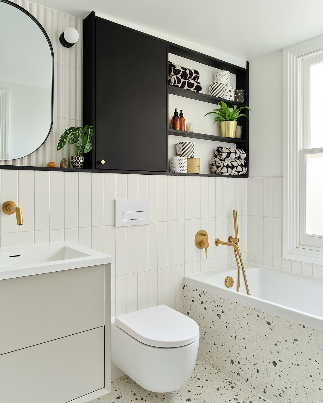 Modern bathroom with white vertical subway tile and terrazzo floor, featuring a black recessed wall unit with a closed cabinet door and open shelving for towels and decor.
