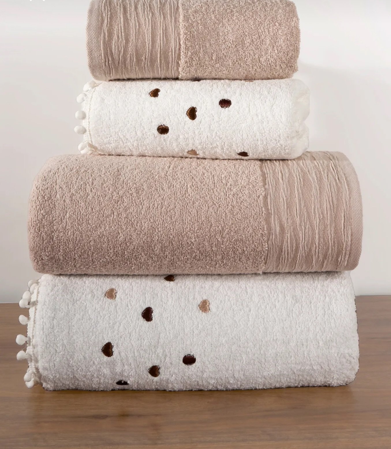 Stack of four alternating towels: solid beige with ridged border, and white with dark heart patterns and pom-pom trim.