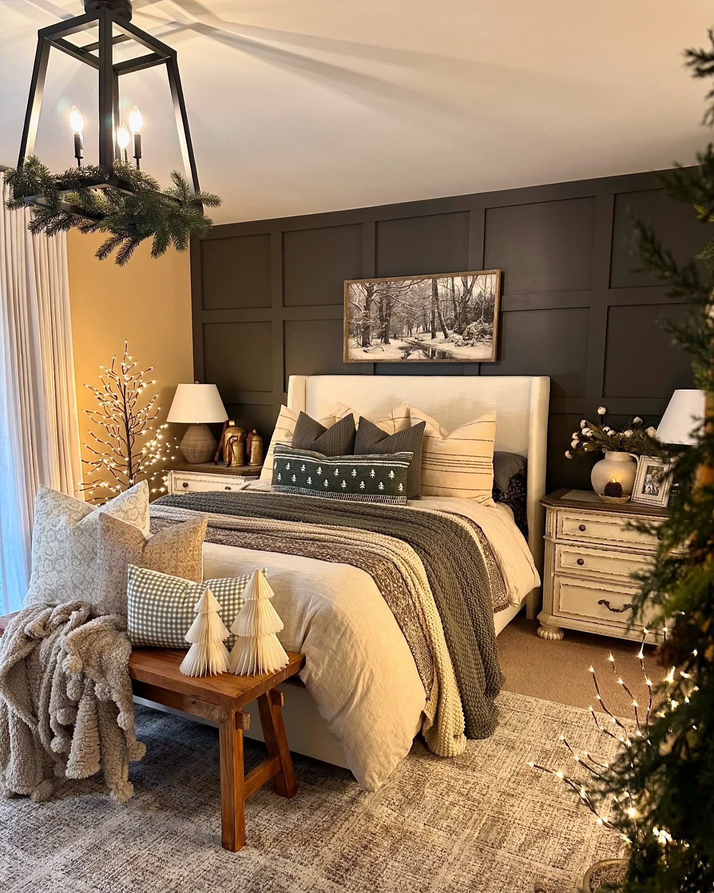 A cozy bedroom featuring a dark gray paneled accent wall, a light upholstered headboard, and layers of bedding in cream, dark gray, and forest green knit, with Christmas accents.