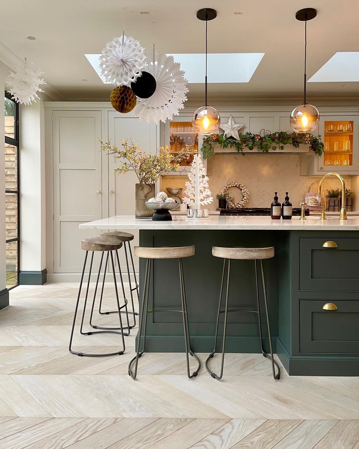 A contemporary kitchen with a dark forest green island and sage green perimeter cabinets. The island area is decorated with hanging white, black, and gold paper fan decorations, while the range hood is minimally adorned with a light green garland.