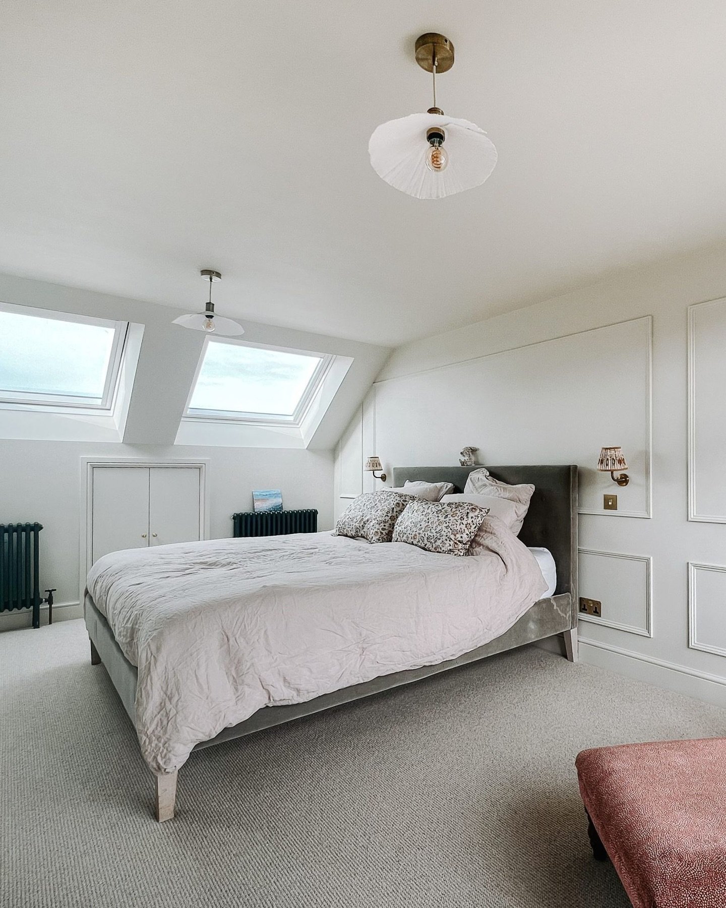A bright and elegant loft bedroom with two skylights, a paneled accent wall, a gray velvet bed, and soft pink bedding.