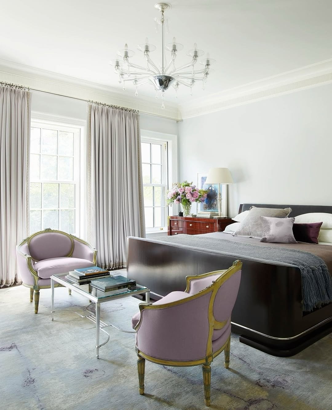 A pair of vintage-style lavender-pink armchairs with gilded wooden frames sits facing each other across a glass table in a luxurious bedroom.