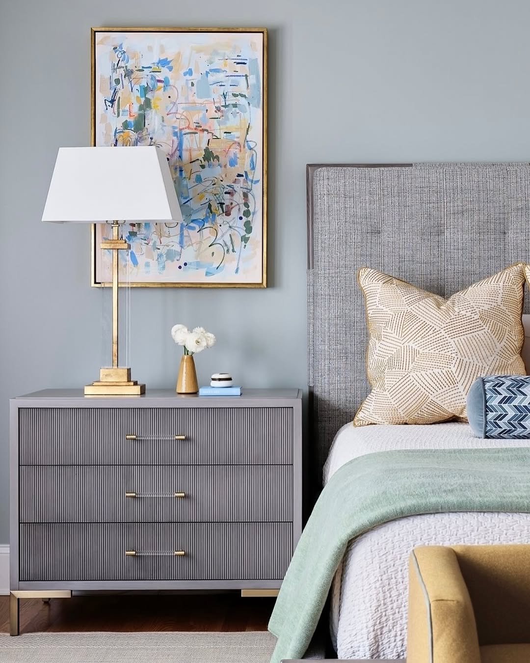 A contemporary gray nightstand with three fluted drawers and thin gold bar pulls. It stands on delicate gold legs. On the top, there is a tall, modern brass lamp with a white rectangular shade and a small vase with white flowers. Abstract art hangs on the wall above.