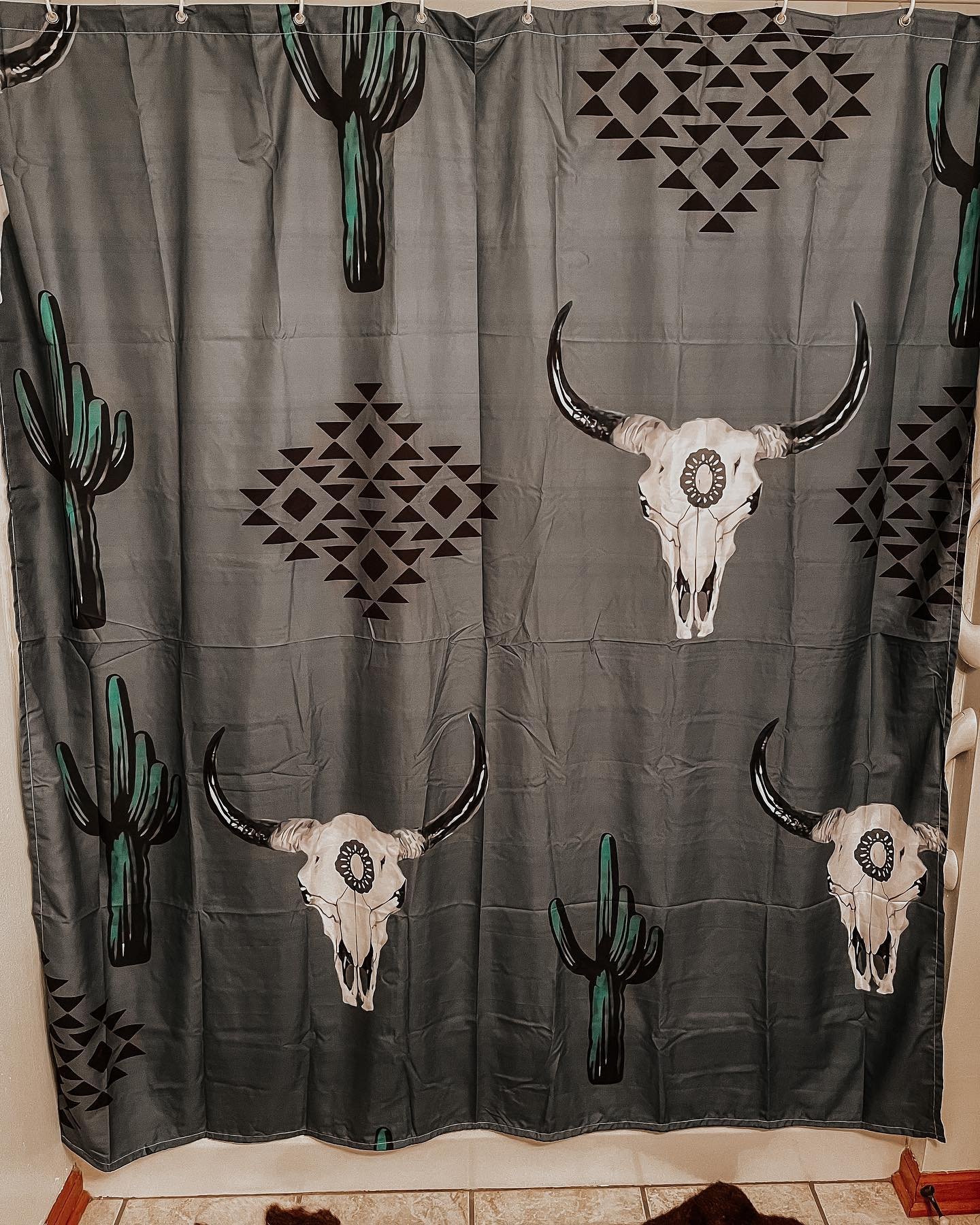 A dark gray shower curtain printed with repeating patterns of white longhorn cow skulls, green cacti, and black geometric Southwestern diamond motifs.