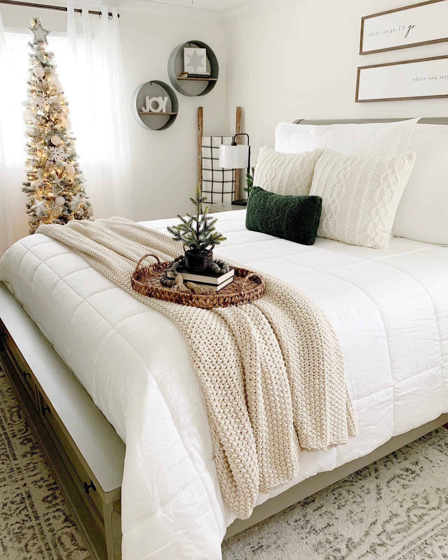 A bright, minimalist bedroom with an all-white bed, a chunky cream knit throw, a pencil Christmas tree, and wall décor featuring stacked circles spelling out "JOY."