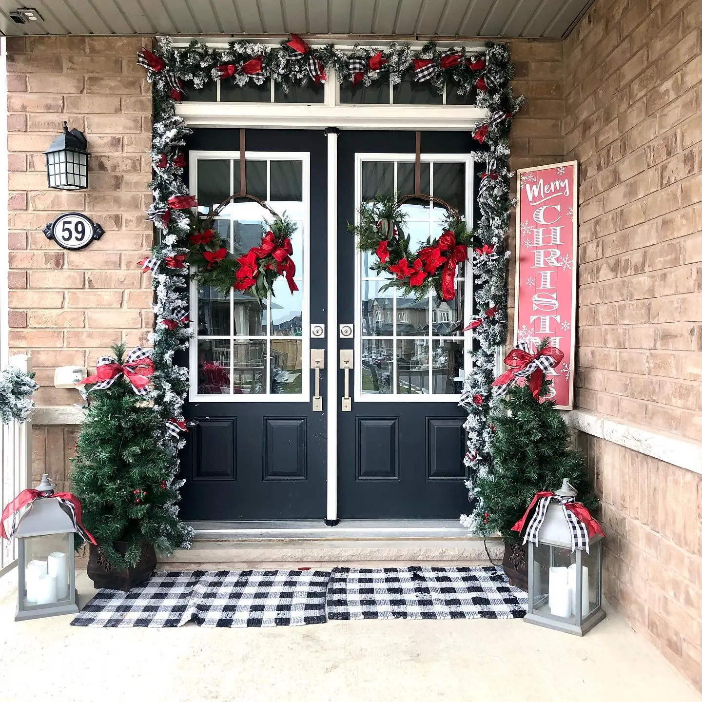 A symmetrical Christmas porch with black double doors, flanked by planters with snow-dusted faux trees and large grey lanterns, with garland and wreaths decorated in red and black gingham ribbon.