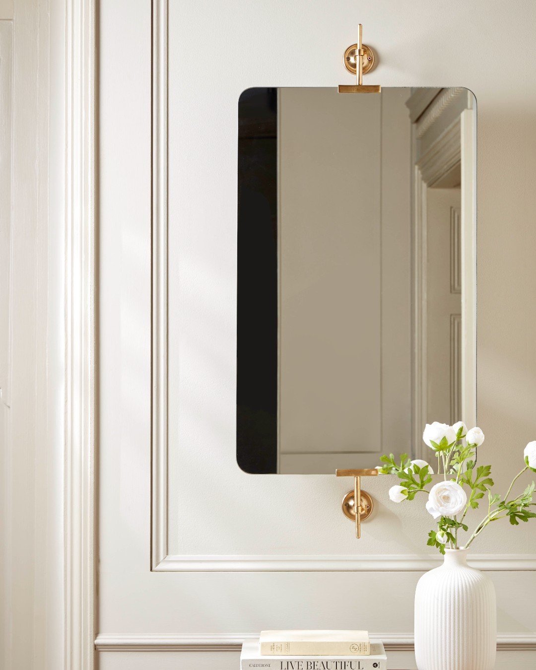 A rectangular wall mirror with rounded corners is mounted on a light-colored wall with two brass brackets.