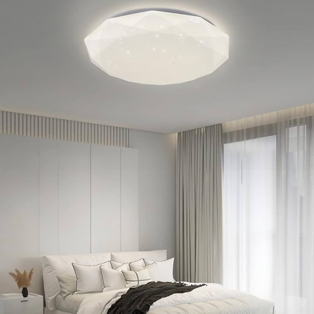 A modern, faceted hexagonal flush mount ceiling light with a starry effect hangs over a white bed in a minimalist bedroom.