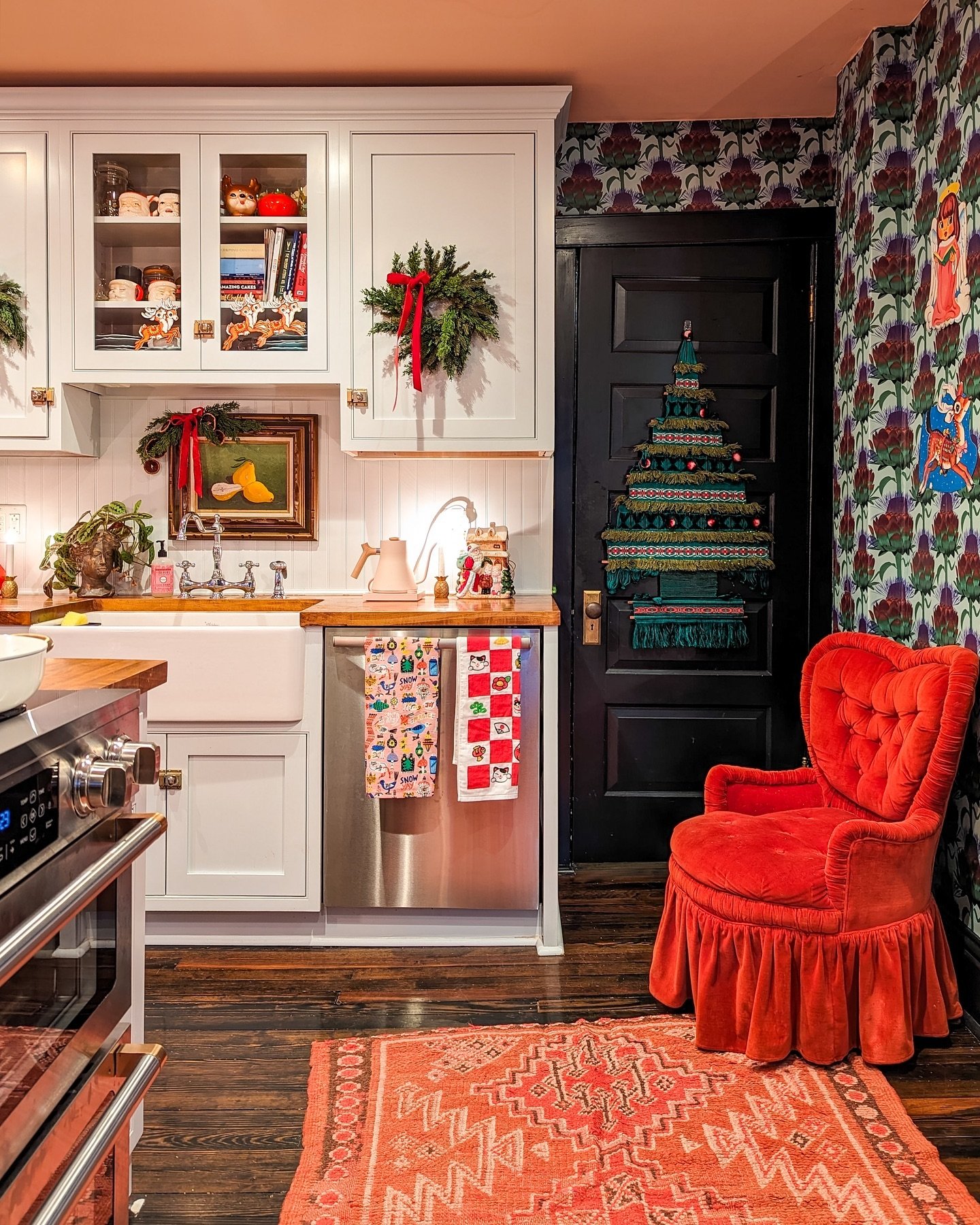 An eclectic kitchen with white shaker cabinets, butcher block counters, and bold floral wallpaper. A vibrant orange-red velvet armchair sits next to a black door decorated with a unique tiered macrame Christmas tree tapestry.