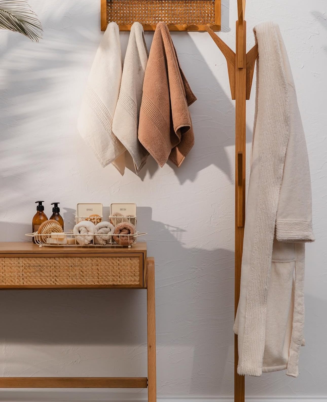 Three earth-toned hand towels hanging from a rattan hook rack next to a cream robe on a wooden stand.