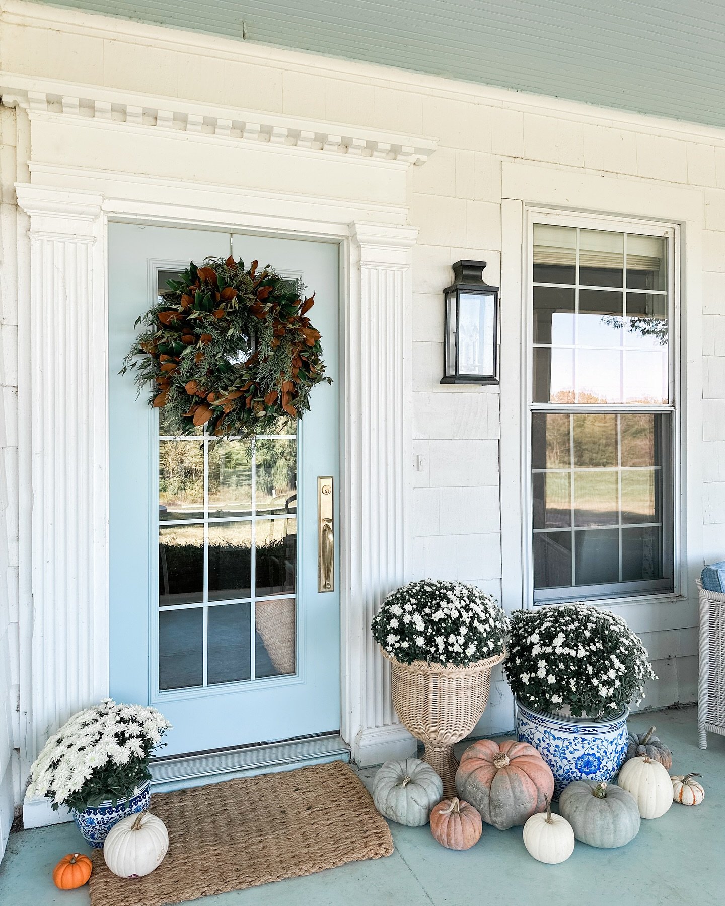 A fall porch decorated with pumpkins, white mums in blue and white planters, and a large wreath on a light blue door.
