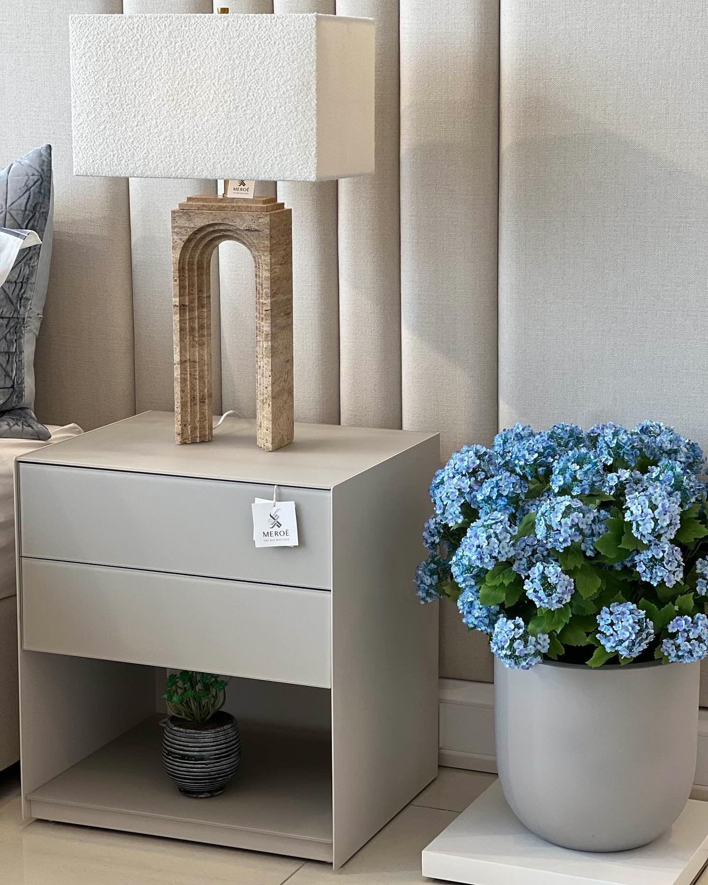 A sleek, modern gray nightstand with two drawers and an open bottom shelf. On top sits a unique, sculptural lamp with an arch design and a white rectangular shade. A large pot of blue flowers sits on a small white stool next to the nightstand.