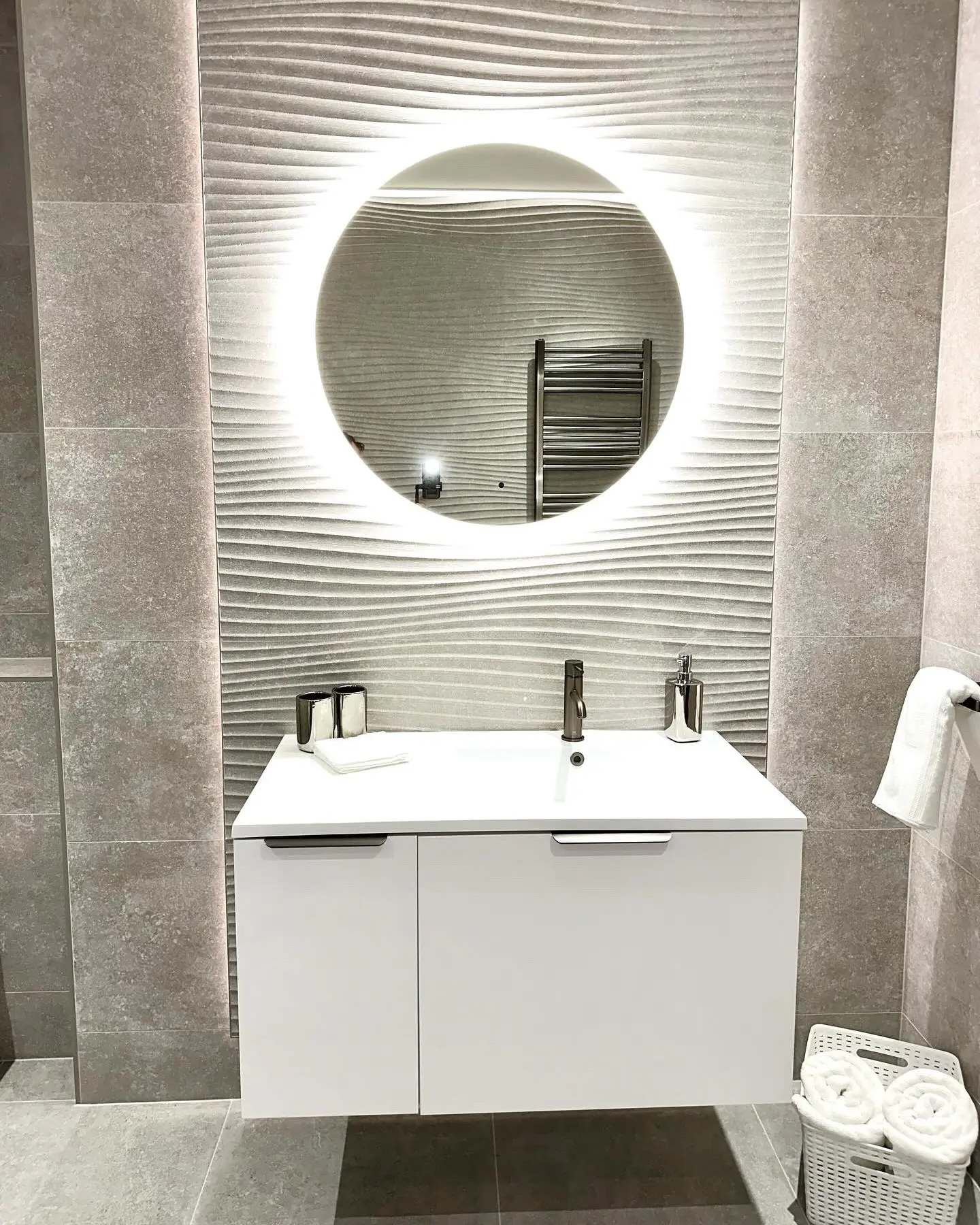 A small white floating bathroom vanity with a square sink and a black faucet, placed against a textured gray wave-patterned wall. A large round backlit LED mirror hangs above the vanity, illuminating the space.