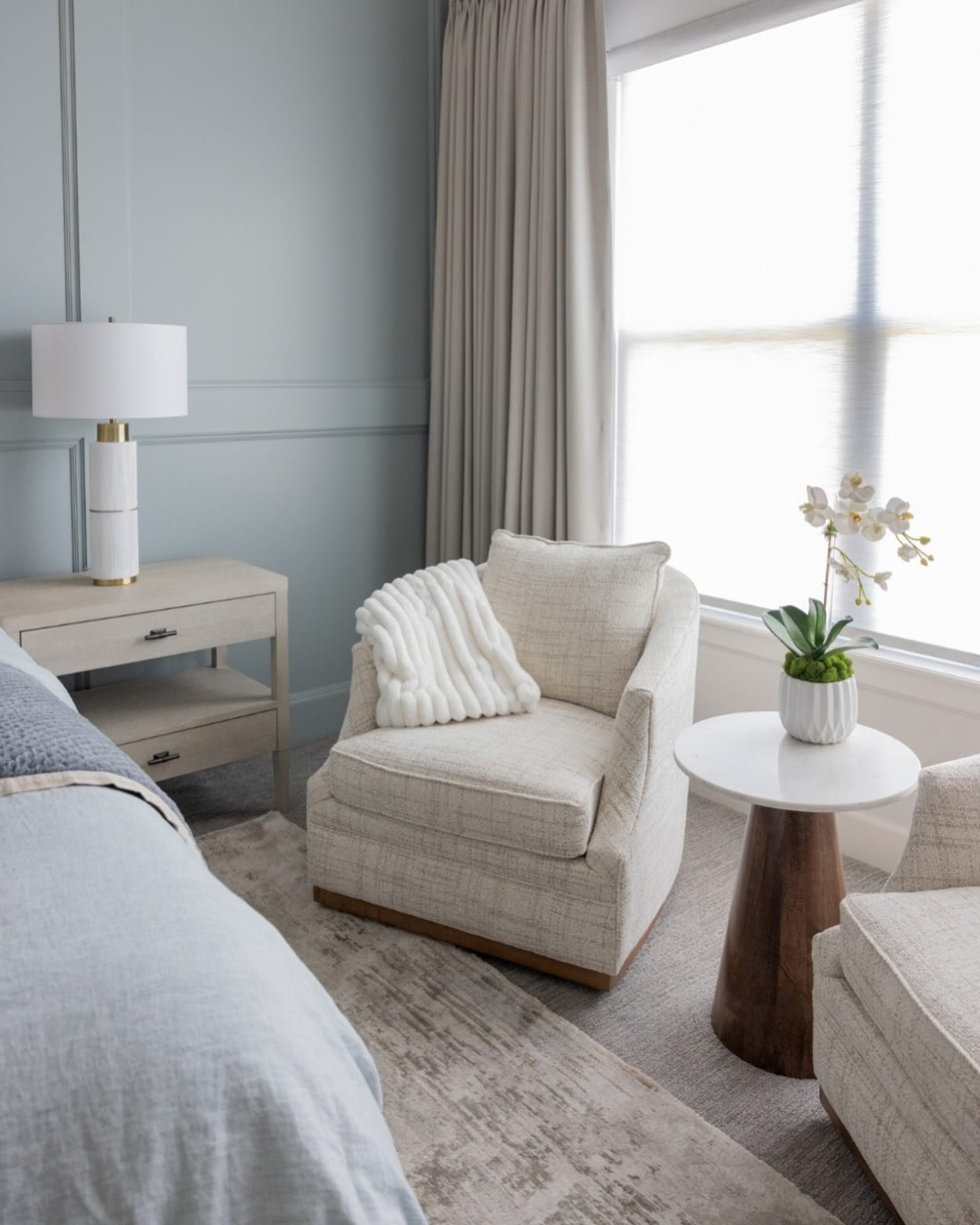 A low, textured club chair with a wooden base and a white throw blanket sits in a sunlit bedroom corner.