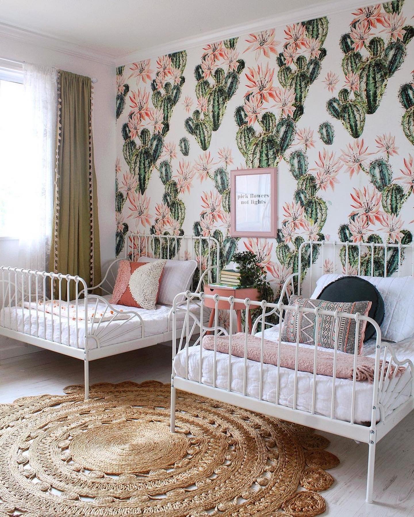 Twin bedroom with cactus-themed wallpaper, white metal beds, a circular jute rug, and soft pink and green accents.