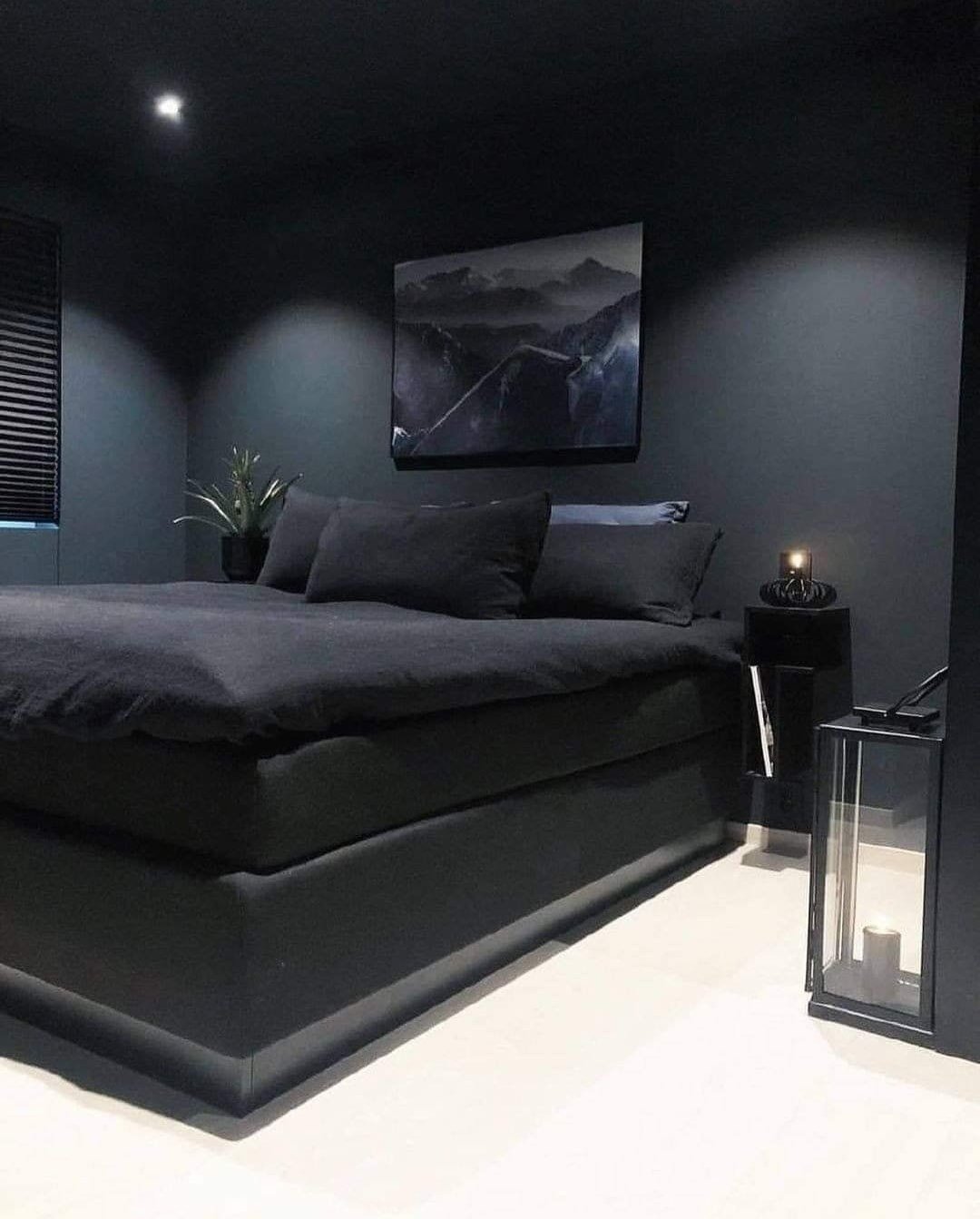 A dark bedroom with matte black walls and ceiling, a large black bed, a single piece of moody landscape art, and a floor lantern.