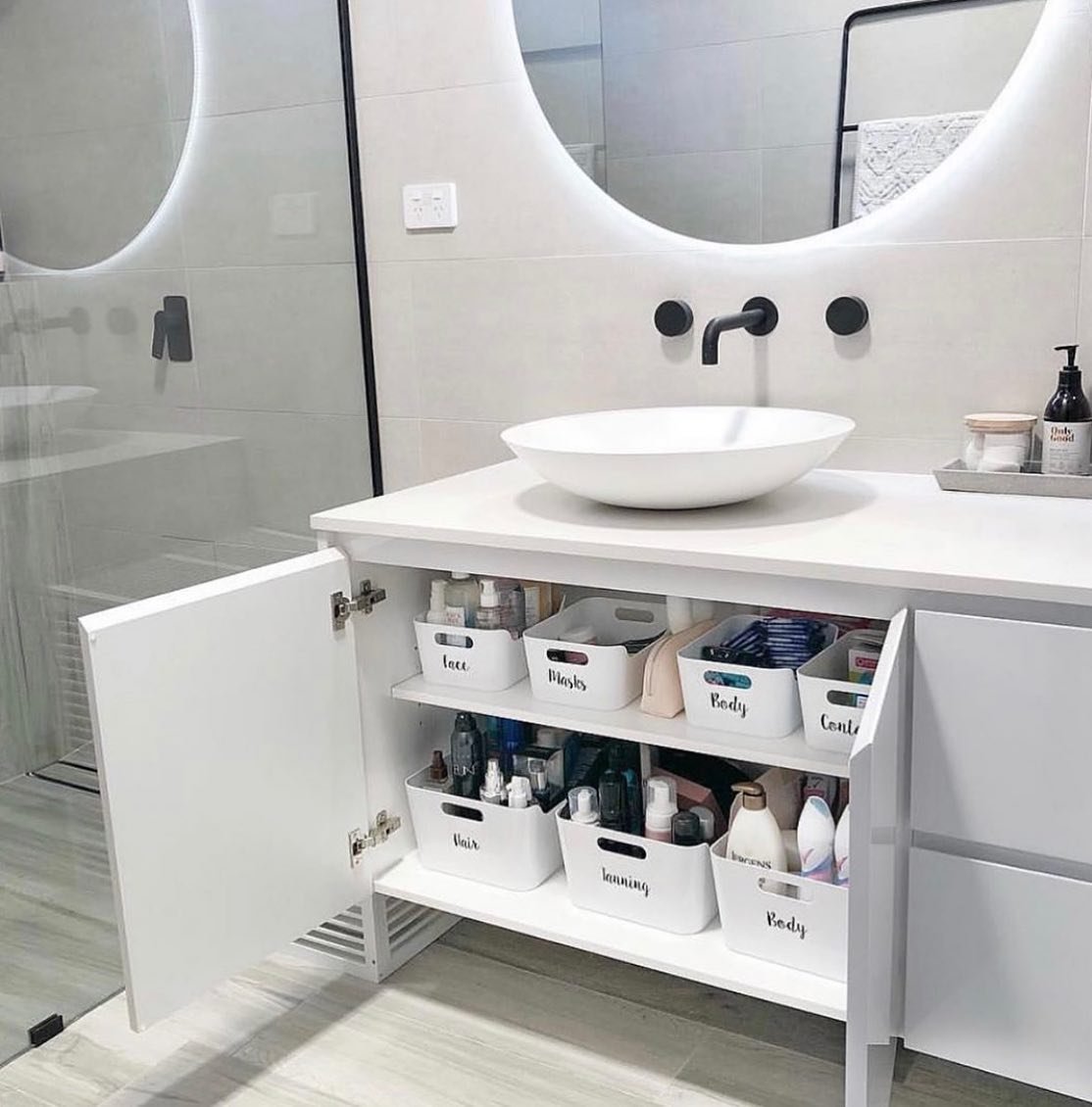 White under-sink cabinet with the doors open, revealing two internal shelves stacked with labeled white plastic storage bins containing toiletries.